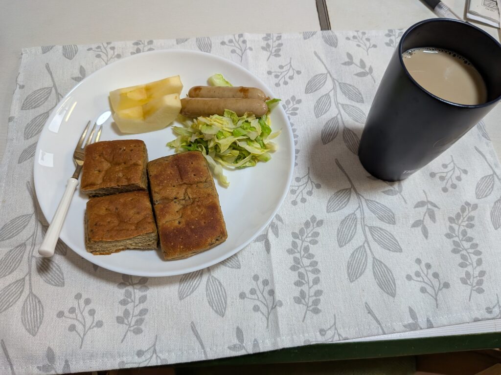 40日目 朝食:白皿に四角い BASE BREAD〈コーヒー味〉2片・炒めキャベツ・鶏ソーセージ1本・りんごくし切り2片が並び、右に黒タンブラーの無糖カフェオレが置かれている。