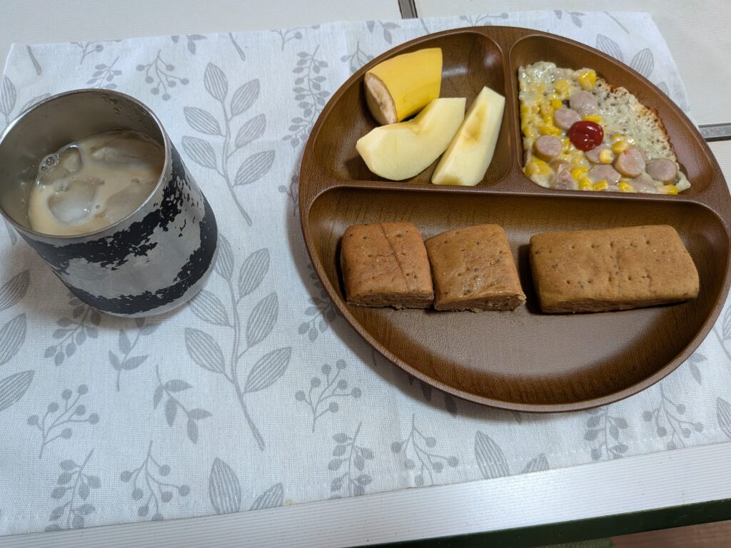 41 日目 朝食:木製プレートに BASE BREAD〈ストロベリー〉(2片)を3つに切り分けたもの・コーン&ソーセージのミニピザ餃子の皮・リンゴくし切り2片・バナナ1/2本が並び、左に氷入り無糖アイスカフェオレ(牛乳50 mL)が置かれている。
